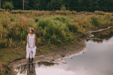 Gün batımında göl kenarında duran güzel boho kızı. Beyaz bohem elbiseli, rüzgarlı saçlı, yeşil yaprakların arasında, suyun yanında dinlenen hasır şapkalı çekici genç bir kadın. yaz tatili