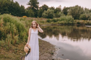 Gün batımında göl kenarında duran güzel boho kızı. Beyaz bohem elbiseli, rüzgarlı saçlı, yeşil yaprakların arasında, suyun yanında dinlenen hasır şapkalı çekici genç bir kadın. yaz tatili