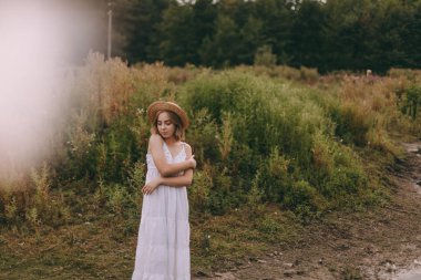 Gün batımında göl kenarında duran güzel boho kızı. Beyaz bohem elbiseli, rüzgarlı saçlı, yeşil yaprakların arasında, suyun yanında dinlenen hasır şapkalı çekici genç bir kadın. yaz tatili