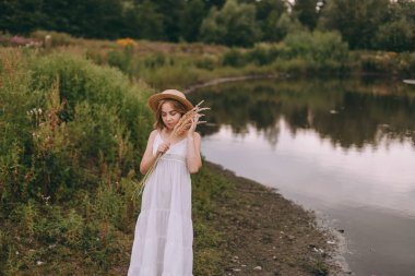 Gün batımında göl kenarında duran güzel boho kızı. Beyaz bohem elbiseli, rüzgarlı saçlı, yeşil yaprakların arasında, suyun yanında dinlenen hasır şapkalı çekici genç bir kadın. yaz tatili