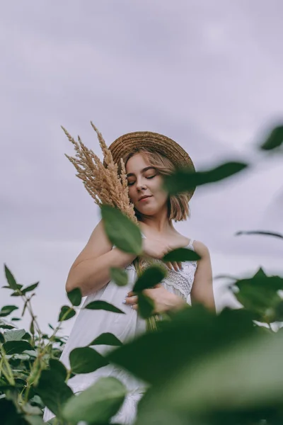 Krásná mladá dívka v bílých šatech a slaměném klobouku v zeleném poli — Stock fotografie