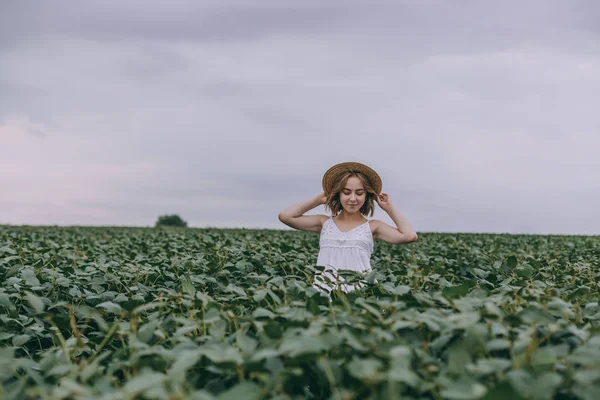 Schönes junges Mädchen in weißem Kleid und Strohhut im grünen Feld umarmt Strauß getrockneter Kräuter — Stockfoto