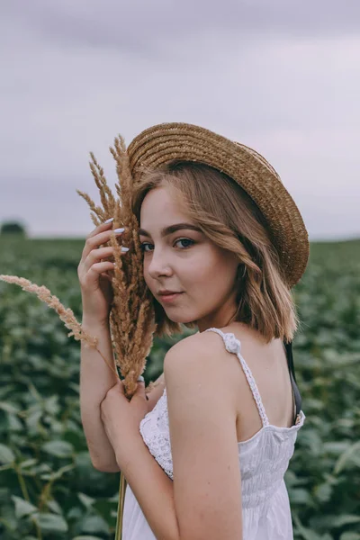 Porträt einer zarten schönen jungen blonden Frau mit strohbraunem Hut in weißem Kleid inmitten grüner Wiesen — Stockfoto