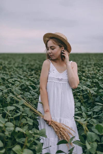 Schönes junges Mädchen in weißem Kleid und Strohhut im grünen Feld umarmt Strauß getrockneter Kräuter — Stockfoto