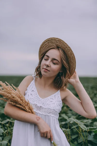 Schönes junges Mädchen in weißem Kleid und Strohhut im grünen Feld umarmt Strauß getrockneter Kräuter — Stockfoto