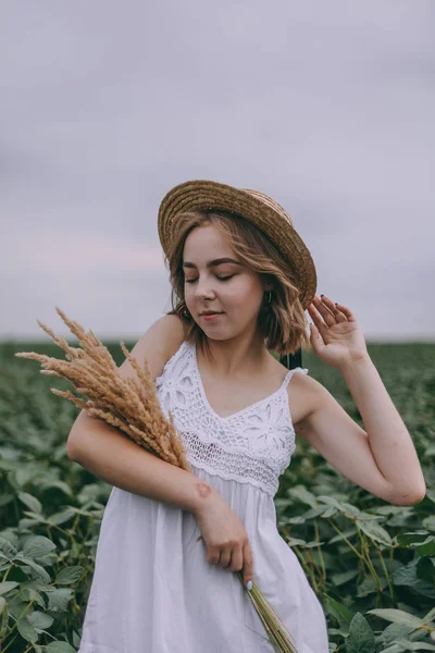 Schönes junges Mädchen in weißem Kleid und Strohhut im grünen Feld umarmt Strauß getrockneter Kräuter — Stockfoto