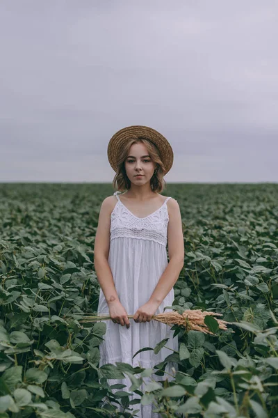 Hermosa joven en vestido blanco y sombrero de paja en campo verde abrazando ramo de hierbas secas —  Fotos de Stock