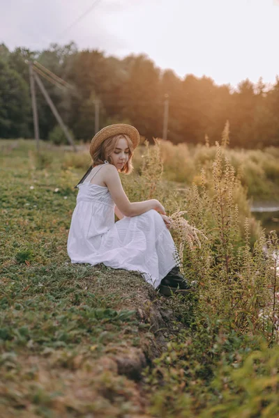 Vacker Boho flicka sitter vid solnedgången ljus nära sjön. attraktiv ung kvinna i vit bohemisk klänning med blåsigt hår avslappnande i närheten av vatten bland gröna blad. Sommarlovet — Stockfoto