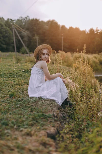 güzel boho kız göl yakınında gün batımı ışıkta oturan. yeşil yaprakları arasında su yakınında rahatlatıcı rüzgarlı saçları ile beyaz bohem elbiseli çekici genç kadın. yaz tatili