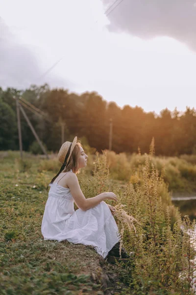 Vacker Boho flicka sitter vid solnedgången ljus nära sjön. attraktiv ung kvinna i vit bohemisk klänning med blåsigt hår avslappnande i närheten av vatten bland gröna blad. Sommarlovet — Stockfoto