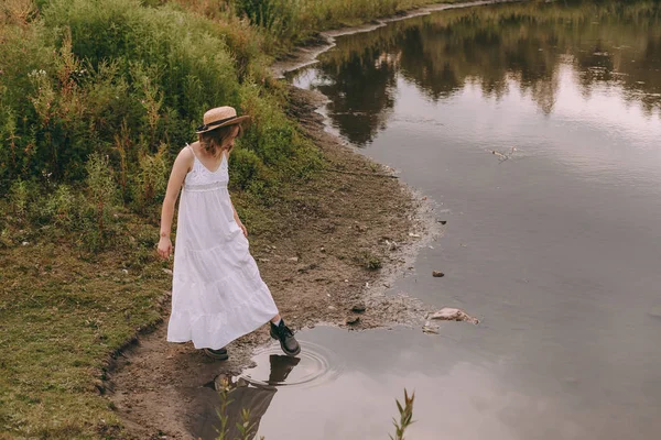 Vacker boho flicka som står vid solnedgången nära sjön. attraktiv ung kvinna i vit bohemisk klänning med blåsigt hår håller halmhatt avkopplande nära vatten bland gröna blad. sommarsemester — Stockfoto
