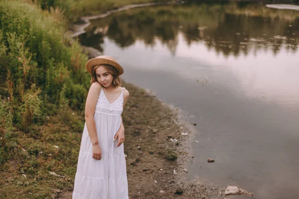 Krásná boho dívka stojí při západu slunce u jezera. atraktivní mladá žena v bílých bohémských šatech s větrnými vlasy drží slamák relaxační u vody mezi zelenými listy. letní dovolená — Stock fotografie