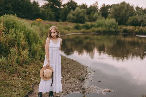 Krásná boho dívka stojí při západu slunce u jezera. atraktivní mladá žena v bílých bohémských šatech s větrnými vlasy drží slamák relaxační u vody mezi zelenými listy. letní dovolená — Stock fotografie