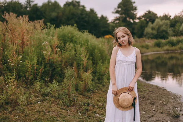 Krásná boho dívka stojí při západu slunce u jezera. atraktivní mladá žena v bílých bohémských šatech s větrnými vlasy drží slamák relaxační u vody mezi zelenými listy. letní dovolená — Stock fotografie