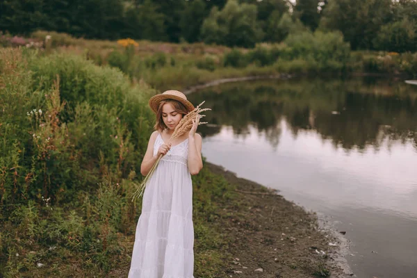 Vacker boho flicka som står vid solnedgången nära sjön. attraktiv ung kvinna i vit bohemisk klänning med blåsigt hår håller halmhatt avkopplande nära vatten bland gröna blad. sommarsemester — Stockfoto