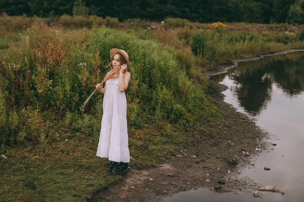 Krásná boho dívka stojí při západu slunce u jezera. atraktivní mladá žena v bílých bohémských šatech s větrnými vlasy drží slamák relaxační u vody mezi zelenými listy. letní dovolená — Stock fotografie