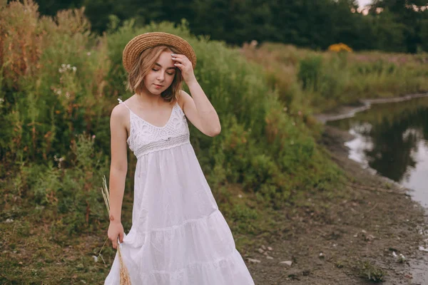 Gün batımında göl kenarında duran güzel boho kızı. Beyaz bohem elbiseli, rüzgarlı saçlı, yeşil yaprakların arasında, suyun yanında dinlenen hasır şapkalı çekici genç bir kadın. yaz tatili