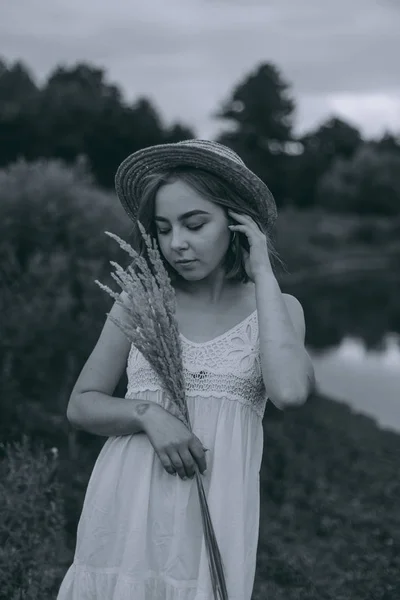Krásná boho dívka stojí při západu slunce u jezera. atraktivní mladá žena v bílých bohémských šatech s větrnými vlasy drží slamák relaxační u vody mezi zelenými listy. letní dovolená — Stock fotografie