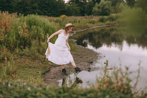 Krásná boho dívka stojí při západu slunce u jezera. atraktivní mladá žena v bílých bohémských šatech s větrnými vlasy drží slamák relaxační u vody mezi zelenými listy. letní dovolená — Stock fotografie