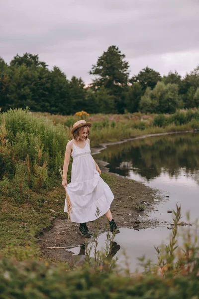 Krásná boho dívka stojí při západu slunce u jezera. atraktivní mladá žena v bílých bohémských šatech s větrnými vlasy drží slamák relaxační u vody mezi zelenými listy. letní dovolená — Stock fotografie