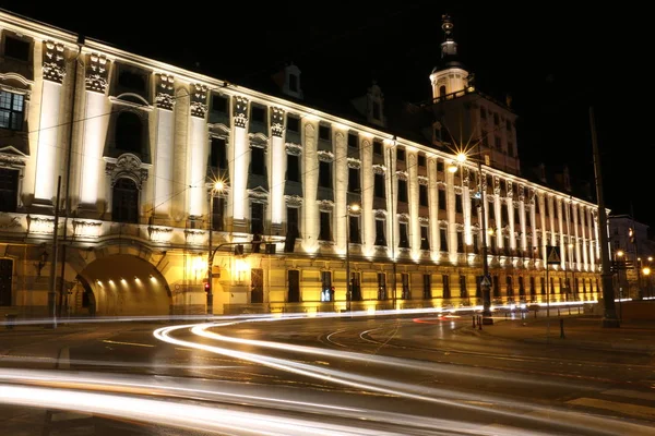 Illuminated main building of the University of Wroclaw at night. A historic building in the Baroque style. Visible representative facade, tower and gate from the Odra river. Streaks of light from cars.