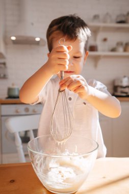 Parlak bir mutfağın arka planında bir çocuk cam bir tabakta pişirmek için hamur yoğurur. Çocuk annesinin yemek pişirmesine yardım eder..