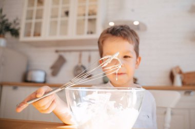 Parlak bir mutfağın arka planında bir çocuk cam bir tabakta pişirmek için hamur yoğurur. Görüntü, hamur damlatan bir çırpıda yakın plan..