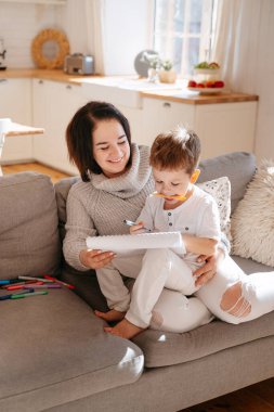 Anne ve oğlu izole bir şekilde gri bir kanepede oturuyorlar. Bir çocukla evde eğitim. Çocuğa yardım et, çocuklarla vakit geçir..
