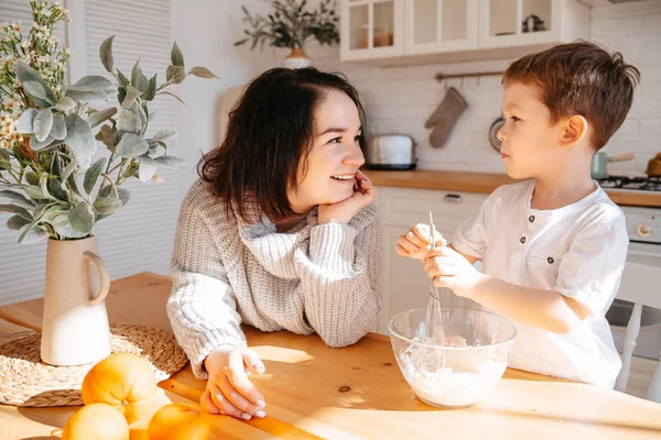 Mom with her son is cooking on the background of a bright kitchen. The child helps parents knead the dough for baking in conditions of self-isolation.The boy is listening to mom.