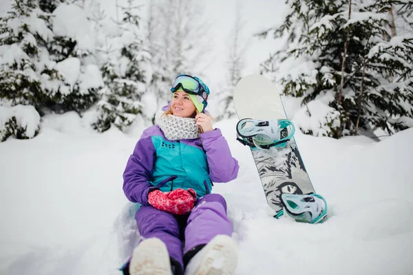 Horizontal. A young girl in sports winter clothes sits on the snow with a snowboard. Happy smiling woman is resting on the mountain.