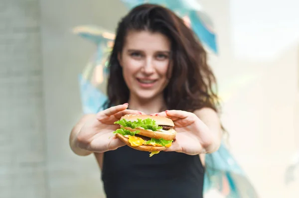 Smiling woman holding burger in hands. Girl Show class Sign. American unhealthy calories meal on background.  Space for text message or design, hungry person smiling with grilled hamburger front view.
