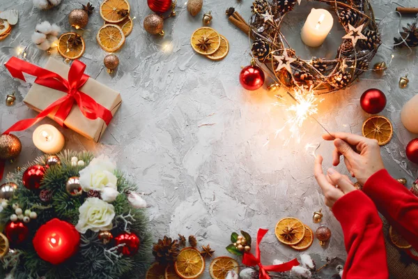 Christmas background with decorations, hands, sparkler, red gift box, orange and light candles. Top view of wreath. Flat lay. Copy space.