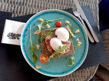 Kavrulmuş pastırmalı yumurta roka, parmesan peyniri dilimleri, vişneli domates ve mavi tabakta kırmızı biberli bezelye.
