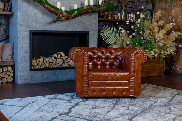 Şöminenin arka planındaki kahverengi deri koltukta yakacak odun, bir buket kurutulmuş çiçek ve mumlar var.