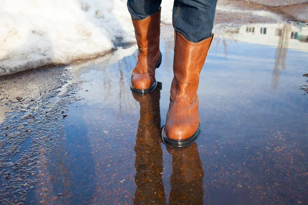 A man in leather boots is on puddles in early spring. Puddles, mud and slush in Russia. Weather and seasons.