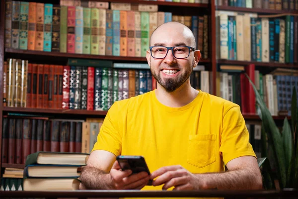 A happy bearded bald man with glasses uses a smartphone. Bookshelves in the background. The concept of a social network, distance learning, and self-isolation.
