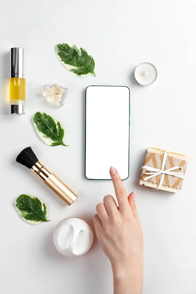 Cosmetics and modern technologies. A woman\'s hand taps the screen of a smartphone, which is lying on a white background with cosmetics and leaves. The concept of organic cosmetics.