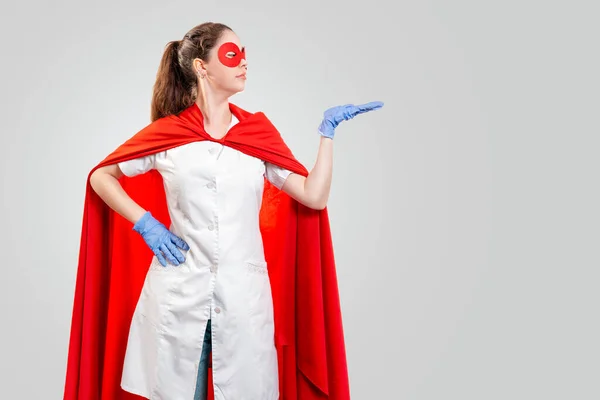 A doctor in a mask, gloves, and superhero Cape points to the side. Gray background. Copy space. The concept of the Power of a super hero for medicine and health care.