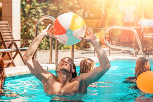 Genç bir atletik adam şişme bir topla havuzda oynuyor. Havuzda yüzen kadınlar ve arka planda plajda güneşlenen kadınlar. Güneş ışığı. Yaz tatili ve arkadaşlık kavramı.