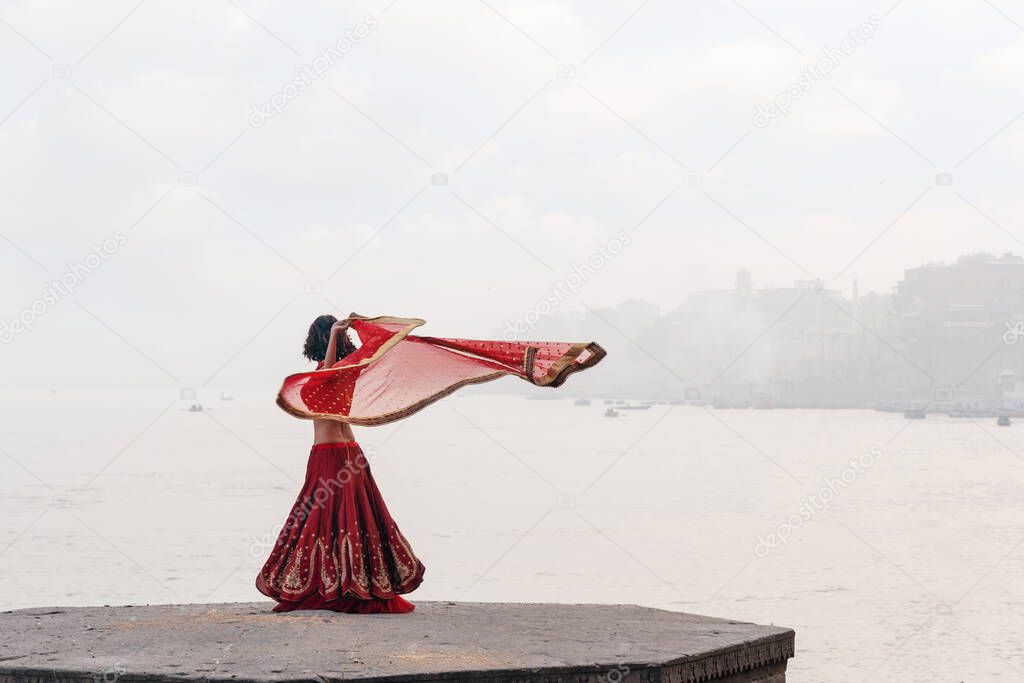 A beautiful Indian woman in a red Sari, dancing with a handkerchief in her hands, alone on the street. In the background, there is a river and a view of the city.