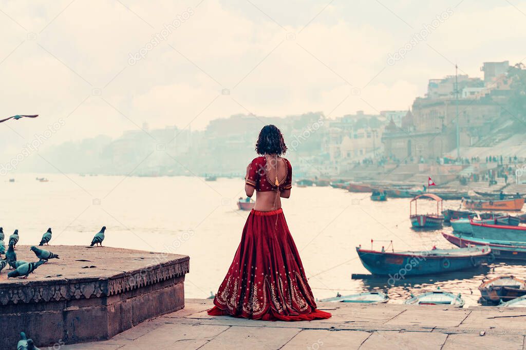 A beautiful Indian woman in a red Sari stands alone on the street. There is a flock of pigeons on the pedestal. In the background is a river and a view of the city. Travel and culture.