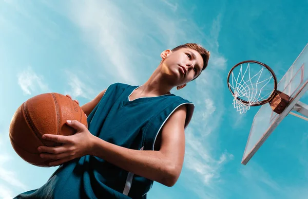 Desporto e basquetebol. Um jovem adolescente em um traje preto fica com uma bola em suas mãos e se prepara para jogar a bola no ringue. Céu azul. Vista inferior — Fotografia de Stock