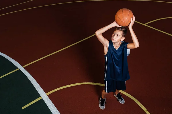 Desporto e basquetebol. Um jovem adolescente de fato de treino azul atira uma bola de salto para o cesto. Terreno no fundo — Fotografia de Stock