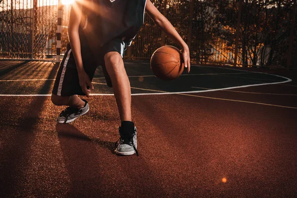 Um jovem adolescente de fato de treino leva uma bola de basquetebol à volta da quadra. Pôr-do-sol ao fundo. Fecha. Conceito de esporte e atividade — Fotografia de Stock
