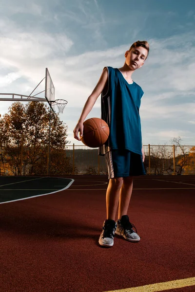Desporto e basquetebol. Um jovem adolescente em um traje preto posa com uma bola de basquete no Parque Infantil. Vertical — Fotografia de Stock