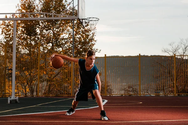 Um jovem adolescente de fato de treino leva uma bola de basquetebol à volta da quadra. Pôr-do-sol ao fundo. Espaço para cópia. Conceito de esporte e atividade — Fotografia de Stock