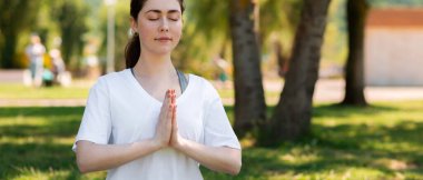 Yoga. Beyaz tişörtlü, gözleri kapalı, çimlerin üzerinde meditasyon yapan genç bir kadının portresi. Uzayı kopyala Panorama. Sağlıklı yaşam tarzı kavramı.