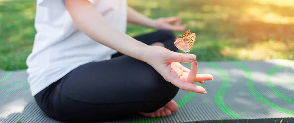 スポーツウェアを着た女性が 芝生の上の公園でマットの上に座って 膝の上に折り畳まれた手で瞑想します サイドビュー パノラマ 蝶のクローズアップで手 リラクゼーションと調和の概念 — ストック写真