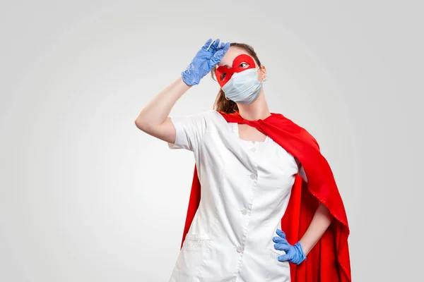 A tired doctor in a medical mask, gloves, and red superhero Cape wipes sweat from her forehead. Gray background. The concept of the Power of a super hero for medicine and stress at work. Copy space.