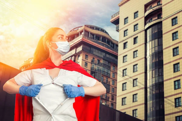 A doctor wearing medical gloves, a surgical mask, and a red superhero Cape rips the medical gown on his chest. Modern buildings in the background. The concept of the Power of a super hero for medicine.